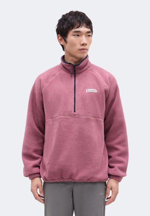 Jeune homme portant un pull polaire violet avec une fermeture éclair partielle, debout devant un fond clair uni, regardant vers la droite.