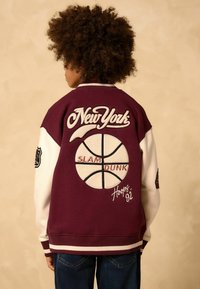 Burgundy színű bomberdzseki fehér ujjakkal, rajta a "New York" felirat és egy kosárlabda grafika. Hímezet és bordás mandzsetta jellemzi.