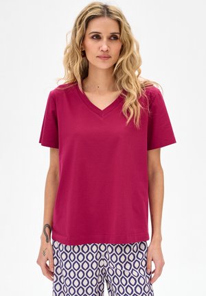 Blondynka o falowanych włosach, ubrana w magentowy top z krótkim rękawem i dekoltem w serek oraz białe spodnie z niebieskimi geometrycznymi wzorami.