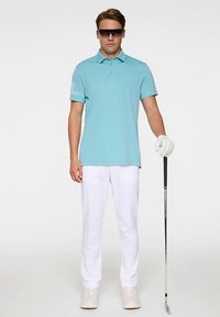 Polo bleu clair à manches courtes avec col et détails décoratifs sur la manche, associé à un pantalon blanc et un club de golf, porté avec un gant.