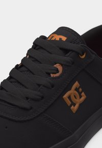 Μαύρο αθλητικό παπούτσι με μαύρα κορδόνια και καφέ λογότυπο DC στο πλάι και στη γλώσσα, με καφέ οπές κορδονιών κοντά στην κορυφή.