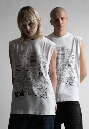 Dvaja mladí dospelí stoja vedľa seba, oblečení v bielych tielkach s čiernym potlačeným textom a abstraktnými vzormi, na jednoduchom sivom pozadí.