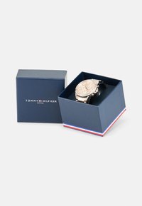 Montre argentée avec un cadran beige, fonction chronographe et bracelet en cuir noir, présentée dans une boîte bleu foncé avec une bordure rouge et blanche.