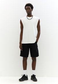Hvid ærmeløs skjorte parret med sorte cargo shorts. Model iført sorte sneakers og hvide sokker, stående mod en ensfarvet baggrund.