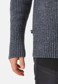 Pull en maille grise à manches longues, présentant une surface texturée. Dispose d'une petite étiquette bleue et jaune sur le côté.