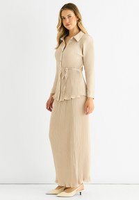 Beige ribbed dress with long sleeves, button front, and a waist tie. Features a ruffled hem. Worn with pointed, light-colored heels.