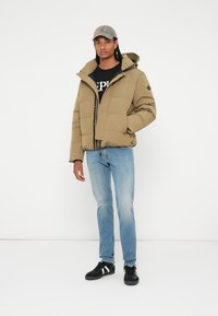 Khaki pufferjakke med hætte og lynlåse, båret over en sort T-shirt. Lyseblå jeans og sorte sneakers med hvide striber.