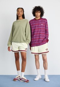 To modeller bærer afslappet sportsbeklædning: en lysegrøn oversized sweatshirt, beige teksturerede shorts med maroon accenter og sko i kontrasterende farver.