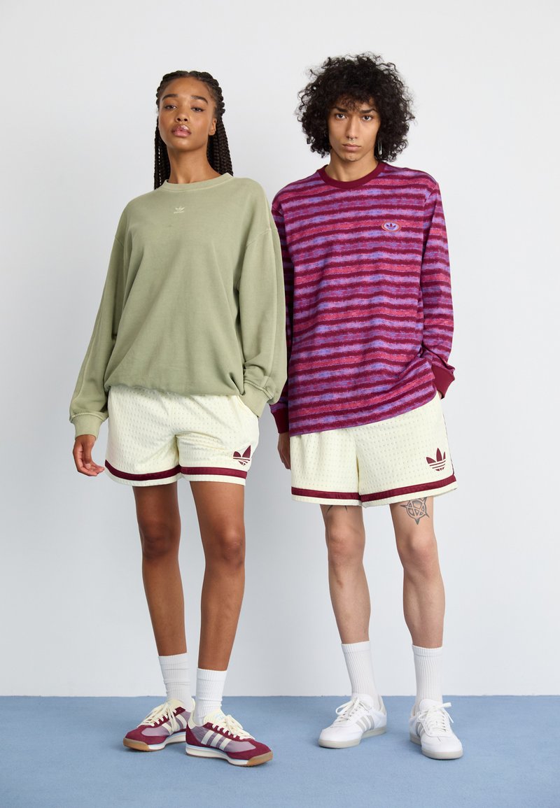 Deux mannequins portent des vêtements de sport décontractés : un sweat-shirt oversized vert clair, un short texturé beige avec des accents bordeaux, et des chaussures de couleurs contrastées.