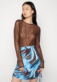 Blusa longa de manga longa transparente castanha com riscas texturizadas, emparelhada com uma saia envolvente de padrão abstrato azul e preto, com laços laterais ajustáveis.