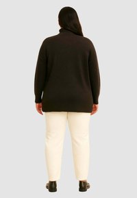 Mørkebrun, blød strikpullover med høj krave, parret med crèmefarvede tætsiddende bukser og sorte slip-on sko.