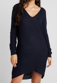 Stickad klänning i marinblått, med V-ringning, långa ärmar och ribbad textur. Sidoklyftor förbättrar rörelse och stil.