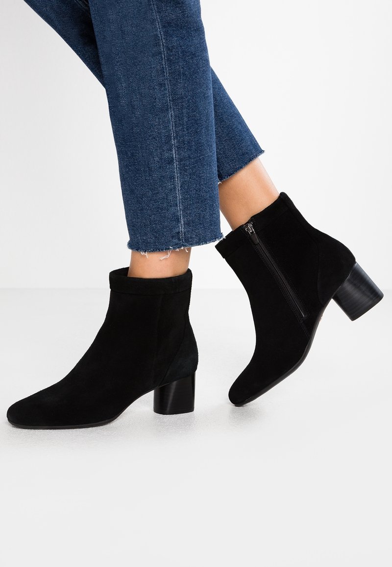 Bottes cheville en daim noir avec un bout rond, un talon bloc de hauteur moyenne et une fermeture éclair latérale. Portées avec un jean bleu raccourci et déchiré.