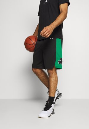 Mand, der holder en basketball, iført sorte og grønne basketballshorts, sort t-shirt, sorte sokker og hvide basketballsko med sorte detaljer.