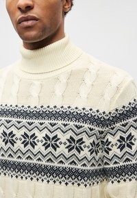 Suéter creme de malha com gola alta, apresentando um padrão geométrico preto e de flocos de neve no peito. Detalhes da textura do tecido visíveis.