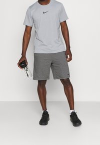 Camiseta gris de Nike con logo negro, combinada con pantalones cortos grises. Zapatillas deportivas negras y una botella de agua transparente en la mano. Diseño sencillo y deportivo.