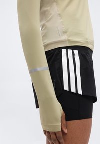 Μακρυμάνικη αθλητική μπλούζα σε ανοιχτό μπεζ χρώμα με ριμπ υφή, συνδυασμένη με μαύρο σορτς που έχει τρεις κατακόρυφες λευκές ρίγες και ανακλαστικές λεπτομέρειες.