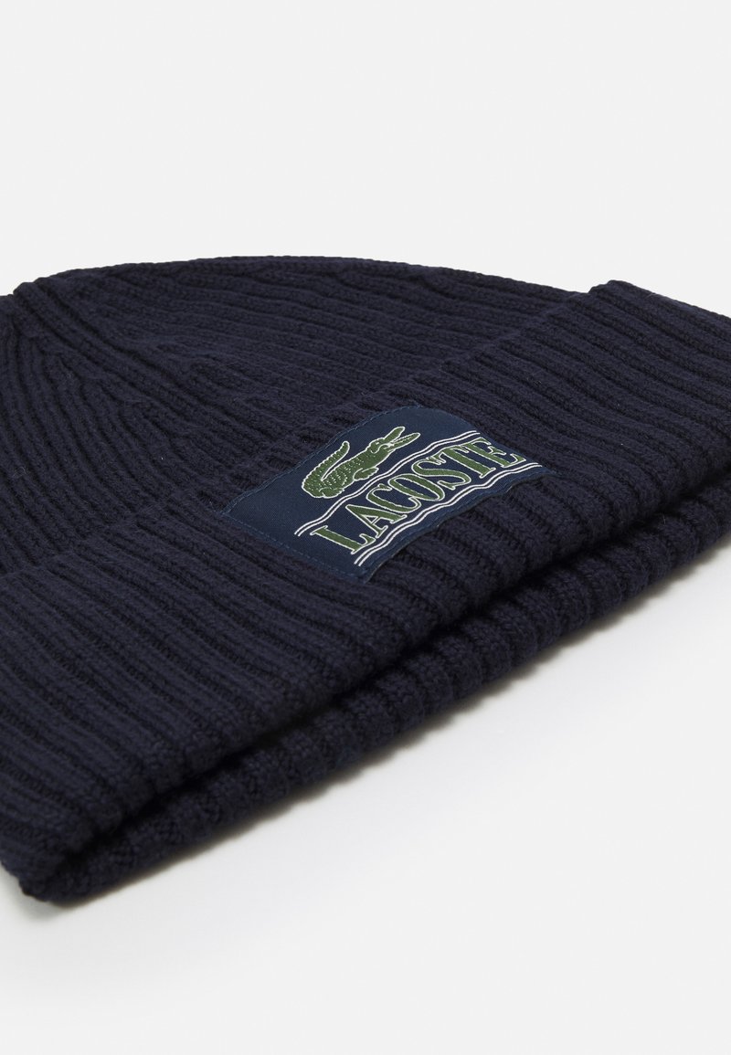 Strickmütze in Marineblau mit geripptem Muster, versehen mit einem grünen gestickten Krokodil-Logo und dem Schriftzug "LACOSTE" auf einem dunklen Patch.
