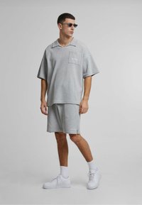 Grå strikket oversized T-skjorte med krage og lomme, kombinert med matchende shorts. Fullført med hvite sneakers og solbriller.