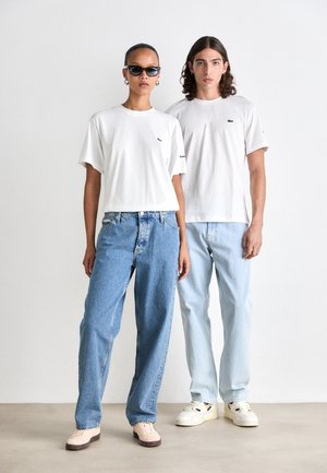 Två modeller står sida vid sida iförda vita Lacoste T-shirts, lösa blåjeans och ljusa sneakers, en av dem bär solglasögon och hoops.