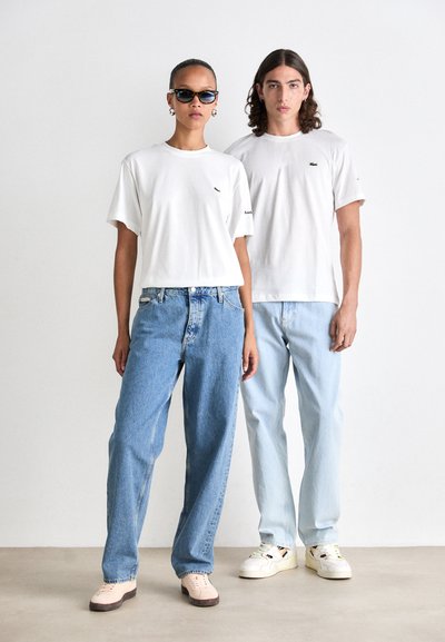 Две модели стоят рамо до рамо, облечени в бели тениски Lacoste, широки сини дънки и светли маратонки, като едната носи слънчеви очила и обеци тип халки.