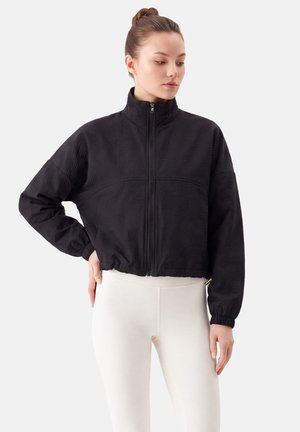 Young woman with hair in a bun wearing a black zip-up jacket and white leggings standing with one hand on her hip against a plain background.