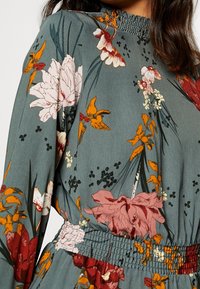 Abito floreale in tessuto teal smorzato con fiori rossi, rosa e gialli. Presenta un collo alto arricciato e maniche lunghe, con una vita aderente.
