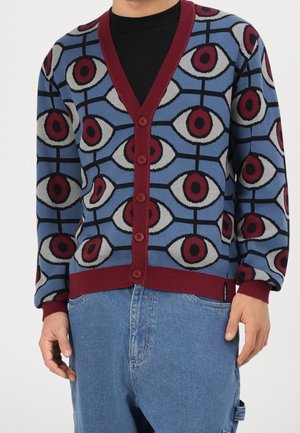 Modrý kardigan se vzorem s výrazným motivem oka v červené a bílé barvě, s výstřihem do V, zapínáním na knoflíky a bordó žebrovanými manžetami a lemem.