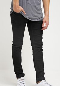 Person som bär svarta slim-fit-byxor, en randig skjorta med handen i fickan, och vita sneakers mot en helt vit bakgrund.