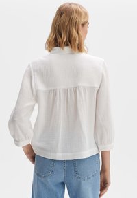 Camicia bianca, tessuto texturizzato, a maniche lunghe con vestibilità ampia, dettaglio arricciato sul retro e colletto semplice, abbinata a jeans azzurri chiari.