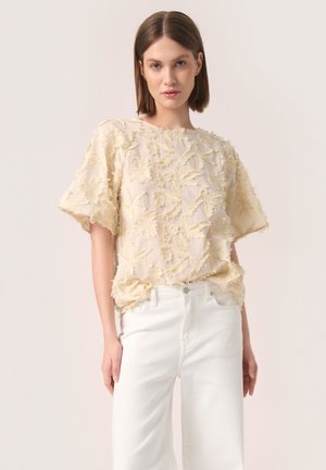 Cremfarbene Bluse mit kurzen Ärmeln und strukturiertem Blumenstickerei, lockere Passform, rundem Ausschnitt, kombiniert mit hoch geschnittenen weißen Jeans.