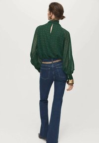 Blusa verde con estampado, de mangas largas y abullonadas, con un corte en forma de llave en la parte posterior, combinada con jeans oscuros de campana y un cinturón dorado como acento.