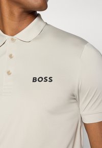 Bež polo majica od glatkog materijala s tri dugmeta, kratkim rukavima i velikim crnim logotipom "BOSS" na prsima.