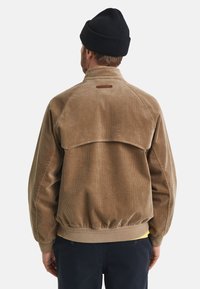 Homem com um casaco de veludo cotelê castanho, um gorro preto e calças escuras de costas contra um fundo branco simples.