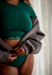 Person iført grønne Calvin Klein undertøj og en grå lynlåstrøje, vist midt i at trække lynlåsen op, stående nær et vindue med lyse gardiner.