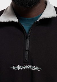 Mustra musteri poolrihmaga must pullover, millel on hall soonikkrae ja rindkere kohal valge ja rohelise tikandiga "ROCAWEAR" logo.