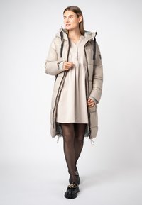 Gevoerde lange jas in licht beige met een capuchon, zwarte accenten en knopen. Gepaard met een beige jurk en zwarte schoenen met een gouden kettingdetail.
