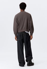 Brauner Sweatshirt mit einem verkürzten Design, kombiniert mit locker sitzenden dunklen Jeans, die sichtbare Gesäßtaschen und gelbe Nähte aufweisen. Schwarze Stiefel.