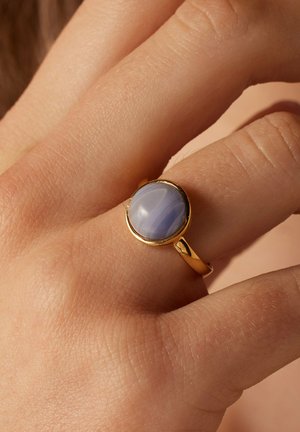 Hand wearing gold ring with round, polished blue lace agate stone on middle finger against beige background.