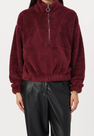 Kobieta w bordowej polarowej bluzie zapinanej na zamek z marszczonym dołem i czarnych skórzanych spodniach, stojąca z rękami swobodnie opartymi po bokach.