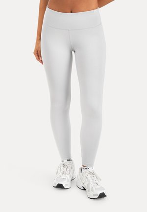 Kvinde iført hvide tætsiddende leggings og hvide sports sko står mod en ensfarvet baggrund.