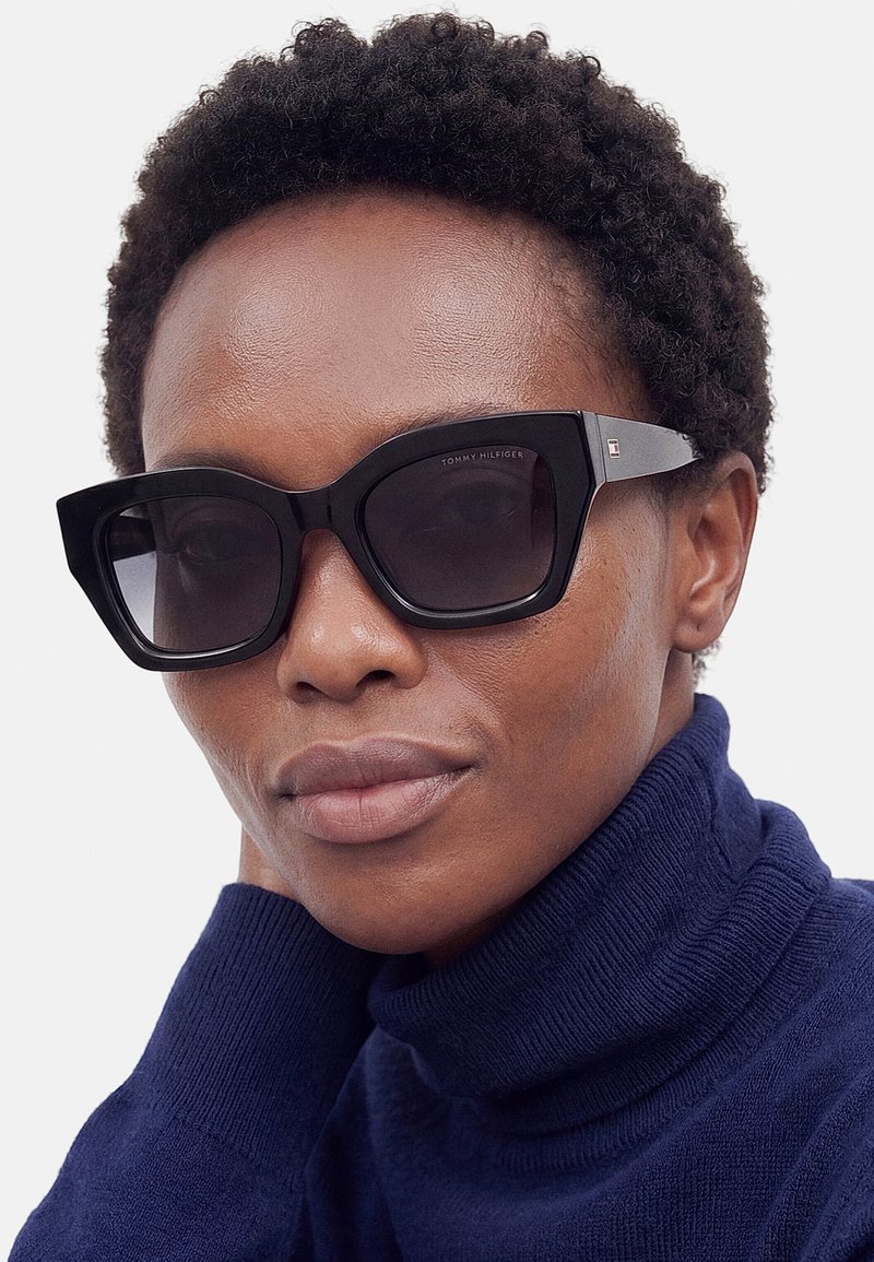 Personne aux cheveux courts et bouclés portant de grandes lunettes de soleil carrées noires et un pull à col roulé bleu foncé, reposant la tête sur la main.