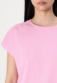 Rožinė marškinėliai su trumpomis rankovėmis ir nėriniuotas apykaklės kraštas. Minkšta medžiaga, apvali iškirptė, lygus paviršius ir laisvas siluetas.
