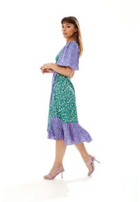 Floral print dress in green and purple, featuring flutter sleeves, a wrap design, and a ruffled hem. Paired with lavender heeled sandals.