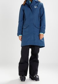 Blauer wasserdichter Parka mit Kapuze, Reißverschluss vorne und zwei Seitentaschen. Wird über schwarzen Skihosen und Stiefeln getragen. Mit Logo auf der Brust.