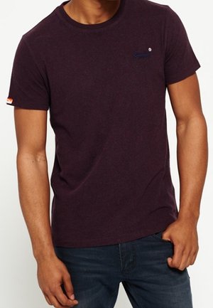 Mand iført en mørkelilla kortærmet t-shirt med et lille logo på brystet og mørkeblå jeans, stående med den ene hånd i lommen.