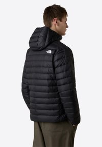 Black puffer jacket with a hood, featuring horizontal quilted sections and a prominent white logo on the left shoulder. Soft, lightweight material.