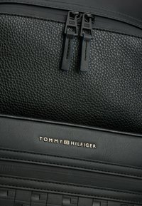 Sac à dos en cuir noir texturé, doté de deux fermetures éclair noires, d'un logo doré, et d'une finition lisse avec un motif près du bas.