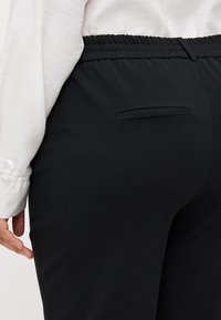 Svarta stretchbyxor med elastisk midja, med en enda bakficka och en slät yta. Närbild som visar tyg och design detaljer.