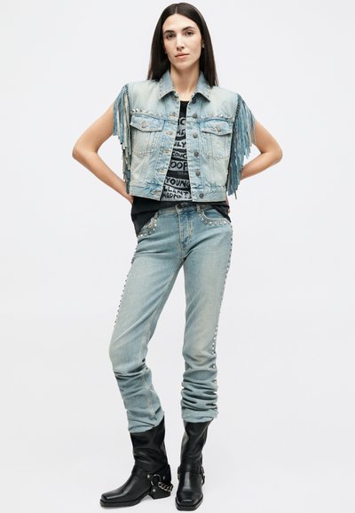 Γυναίκα που φοράει αμάνικο τζιν μπουφάν με φουντίτσες, μαύρο μπλουζάκι με στάμπα, τζιν ανοιχτού μπλε με τρουκς και μαύρες δερμάτινες μπότες, στέκεται με τα χέρια στη μέση.